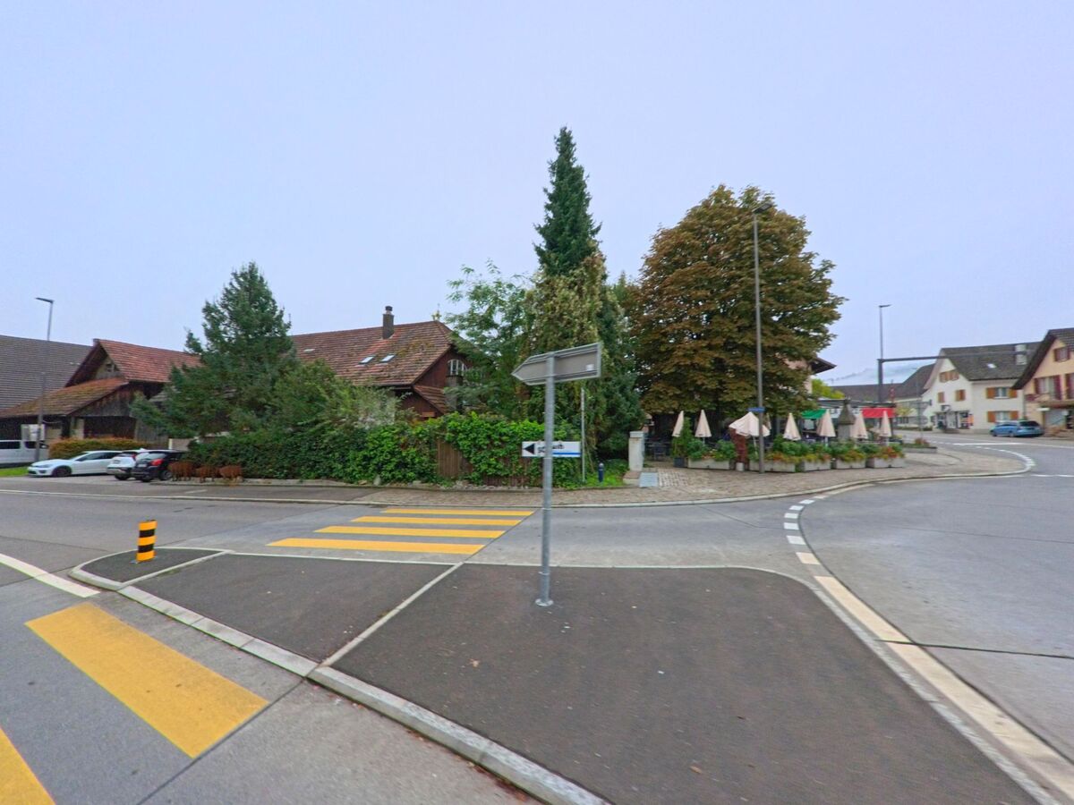 Im Zentrum von Schafisheim (5503, Kanton Aargau) liegt ein kleiner Platz mit Restaurantterrasse und Sonnenschirmen, umgeben von traditionellen Gebäuden.