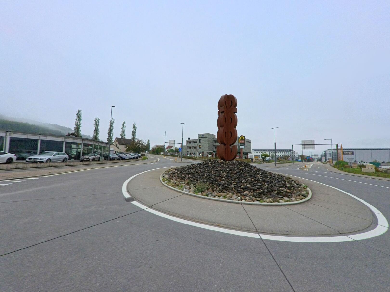 Am Kreisel in Schafisheim (5503, Kanton Aargau) steht eine moderne Metallskulptur auf einem mit Steinen bedeckten Rondell, umgeben von Strassen und Gewerbebauten.