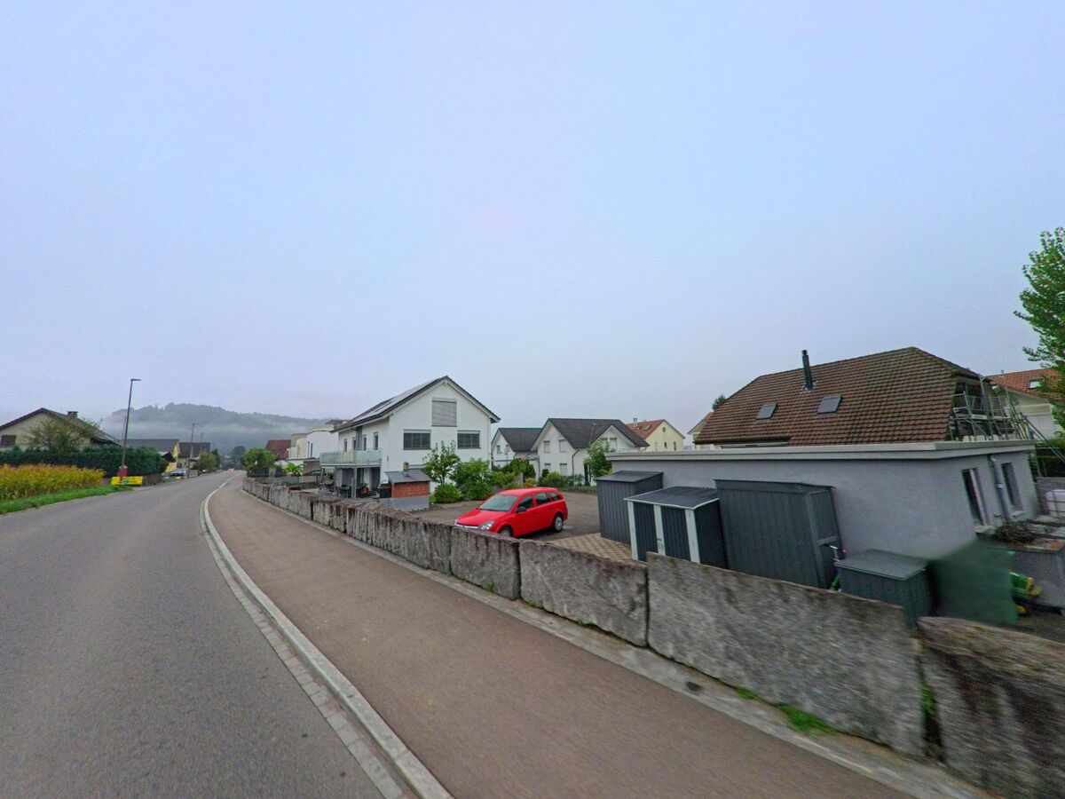 Eine leicht ansteigende Strasse in Schafisheim (5503, Kanton Aargau) führt vorbei an Einfamilienhäusern mit gepflegten Vorgärten und geparkten Autos.