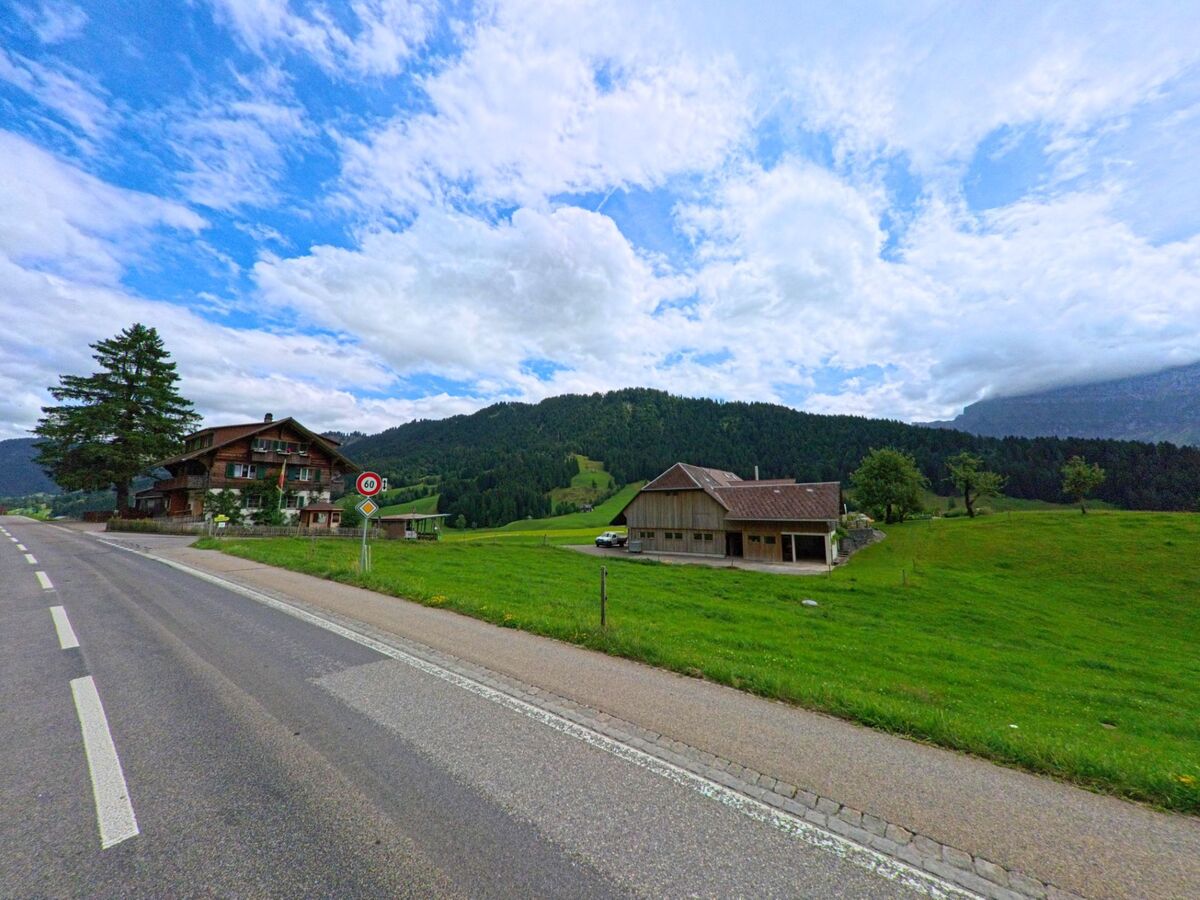 Entlang einer Landstrasse in Schangnau (6197, Kanton Bern) stehen ein Bauernhaus und ein Wohnhaus vor einer bewaldeten Hügellandschaft unter wechselhaftem Himmel.