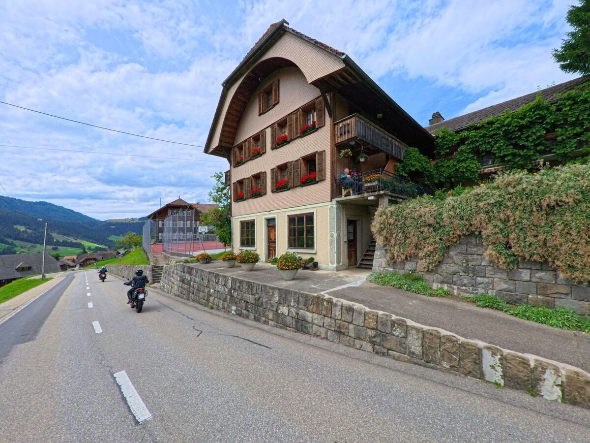 In Schangnau (6197, Kanton Bern) fahren Motorradfahrer an einem traditionellen Haus mit Fensterläden und Balkon vorbei, das erhöht an einer Steinmauer liegt.