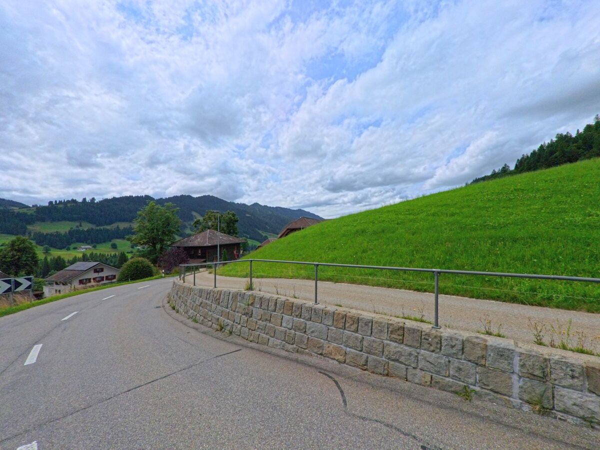In Schangnau (6197, Kanton Bern) verläuft eine kurvige Strasse entlang einer grünen Böschung mit Blick auf verstreute Häuser und bewaldete Hügel.
