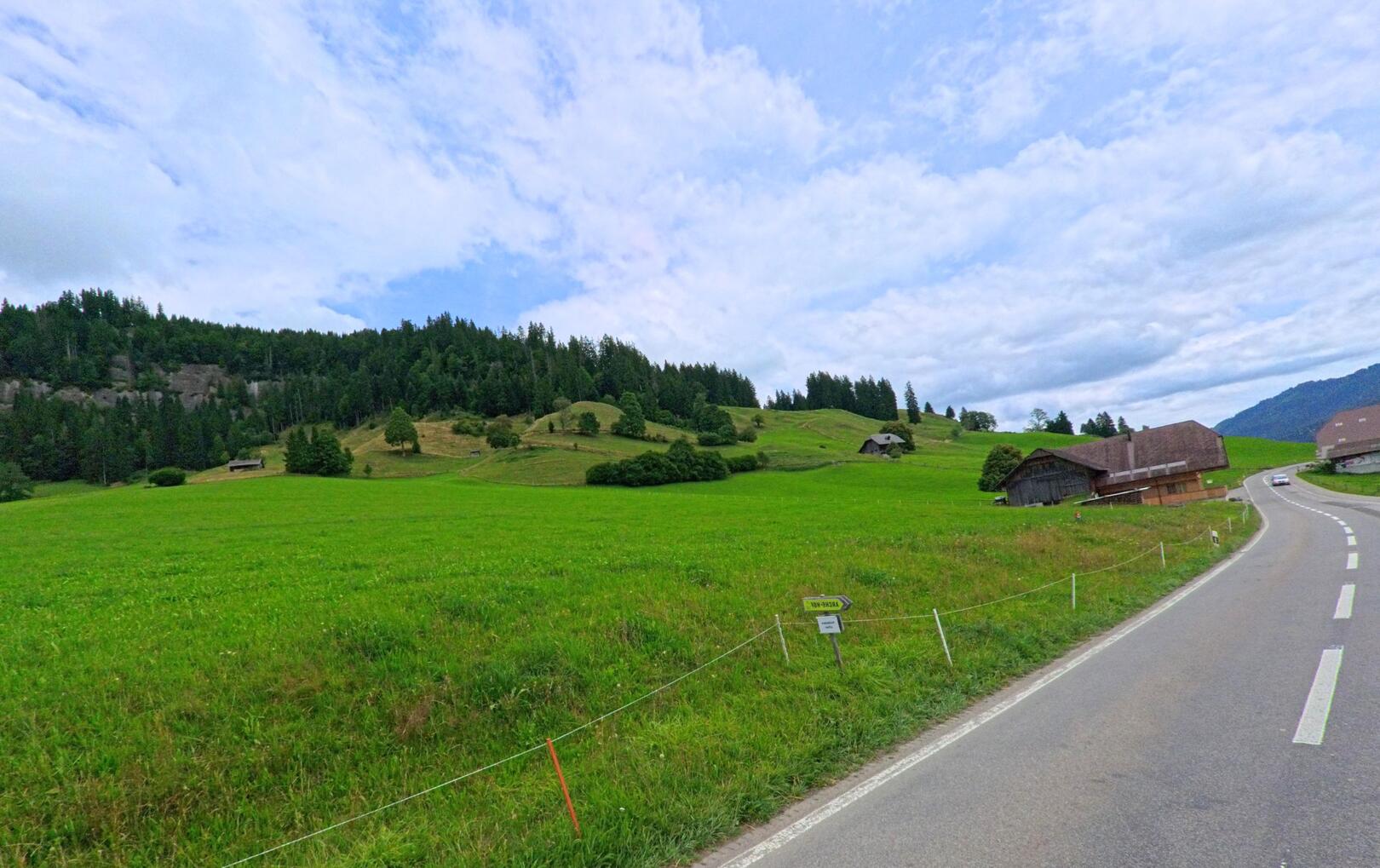 In Schangnau (6197, Kanton Bern) schlängelt sich eine Landstrasse durch grüne Wiesen mit verstreuten Bauernhäusern und bewaldeten Hügeln im Hintergrund.