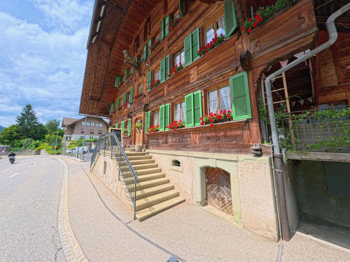 In Schangnau (6197, Kanton Bern) führt eine Treppe zu einem traditionellen Holzhaus mit grünen Fensterläden und roten Blumen direkt an der Strasse.