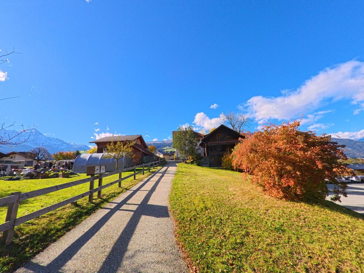 Ein schmaler Weg fuehrt zwischen Gruenflaechen, Zaeunen und Holzbauten durch das Dorfgebiet von Scharans (7412, Kanton Graubuenden) bei herbstlicher Stimmung.