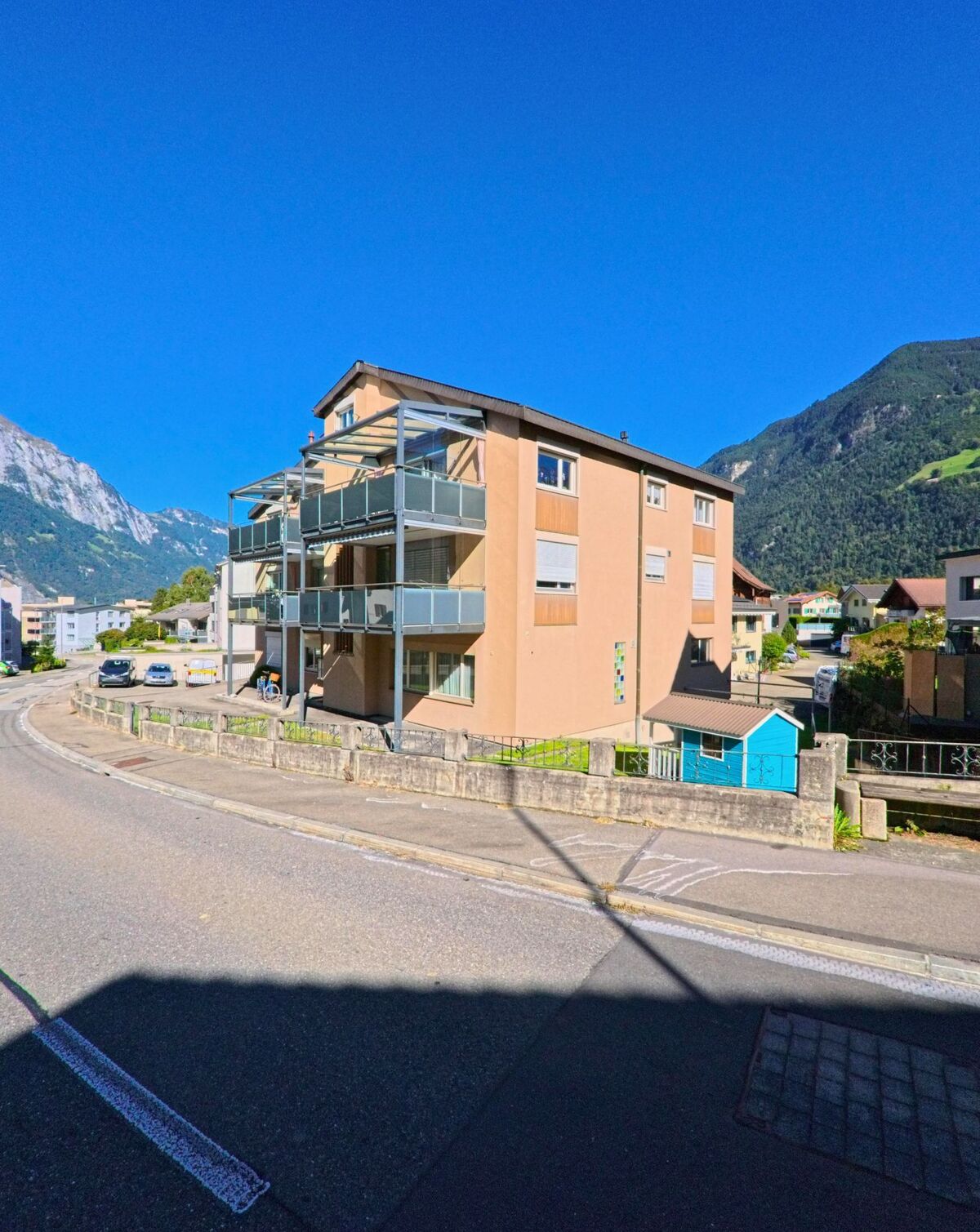 Ein Mehrfamilienhaus mit verglasten Balkonen steht in Schattdorf (6467, Kanton Uri) an einer leicht geschwungenen Strasse mit Blick auf die umliegenden Berge.