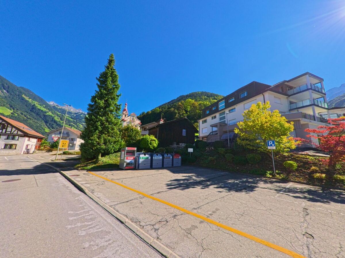 Mehrfamilienhaeuser und Recyclingcontainer stehen in Schattdorf (6467, Kanton Uri) an einer Strasse mit Blick auf die Pfarrkirche und die umliegenden Berge.