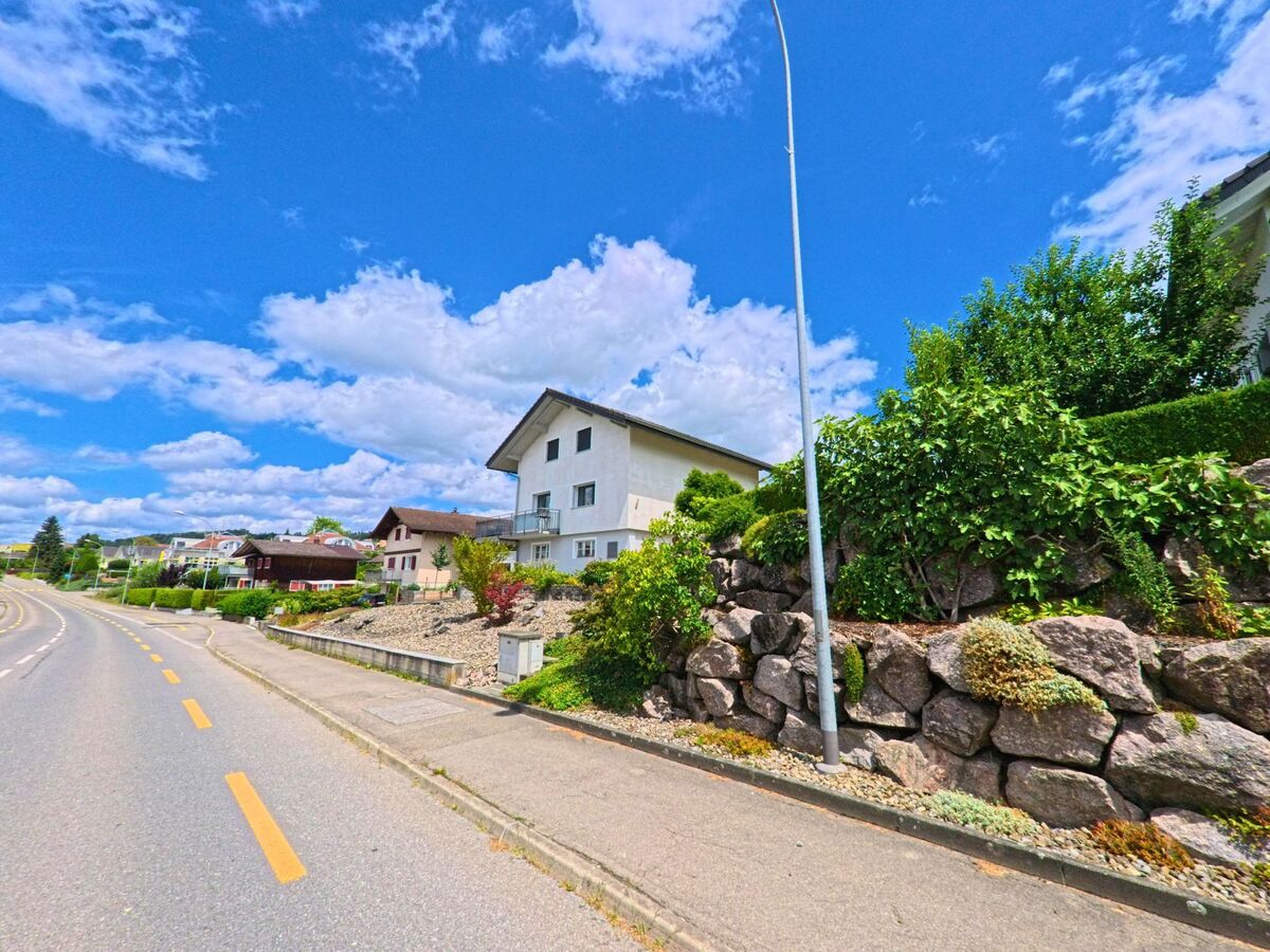 Entlang einer leicht geschwungenen Strasse in Schenkon (6214, Kanton Luzern) stehen Einfamilienhaeuser mit vorgelagerten Stützmauern aus Naturstein und gepflegten Gärten.