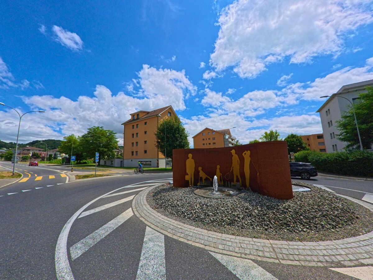 Ein Kreisel in Schenkon (6214, Kanton Luzern) ist mit einer künstlerischen Skulptur und einem kleinen Brunnen in der Mitte gestaltet, umgeben von Wohngebäuden und Strassen.