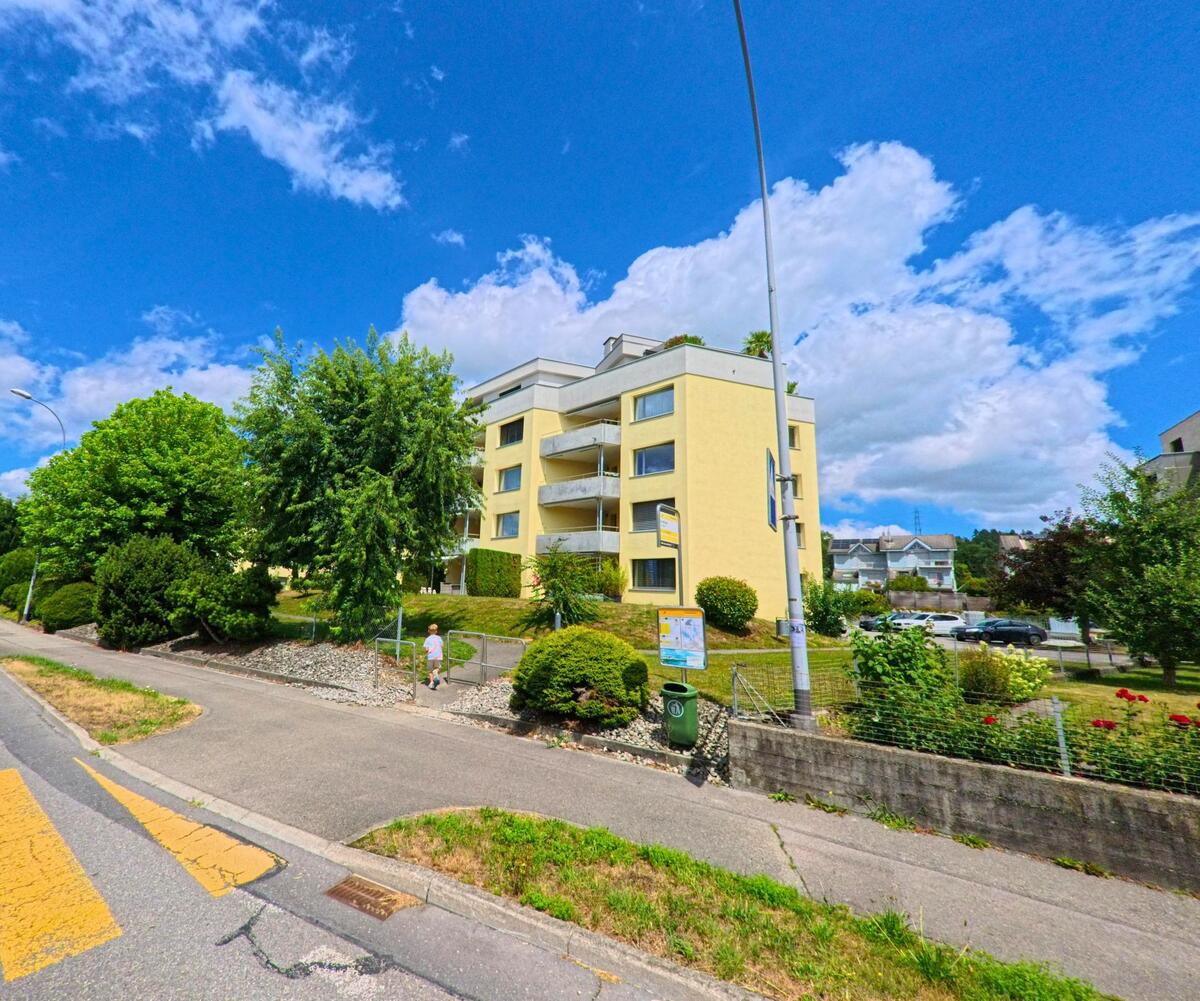 In Schenkon (6214, Kanton Luzern) steht ein gelbes Mehrfamilienhaus mit gepflegtem Vorgarten an einer Strasse mit Fussgängerbereich.