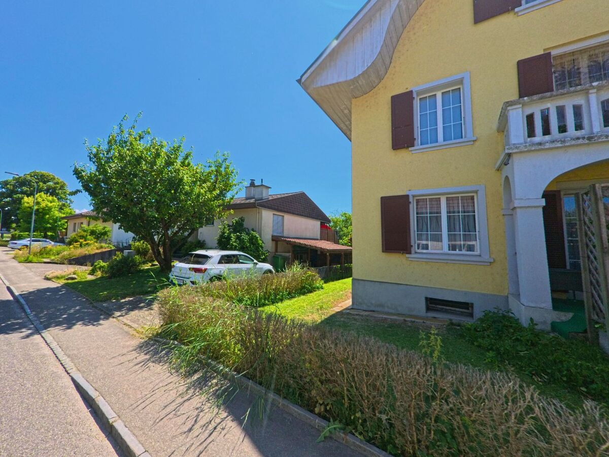 In einem Wohnquartier von Scheuren (2556, Kanton Bern) steht ein gelbes Mehrfamilienhaus mit markantem Dach neben gepflegten Gärten und parkierten Autos.