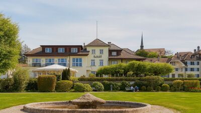 In Murten (3280, Kanton Freiburg) steht das Gebaeude des Hotels Schiff am See, umgeben von gepflegtem Garten und Blick auf einen Brunnen im Vordergrund.