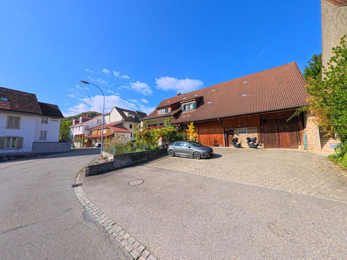 Ein traditionelles Wohnhaus mit grossem Scheunendach und gepflasterter Einfahrt steht an einer Nebenstrasse in Schinznach (5107, Kanton Aargau) bei sonnigem Wetter.