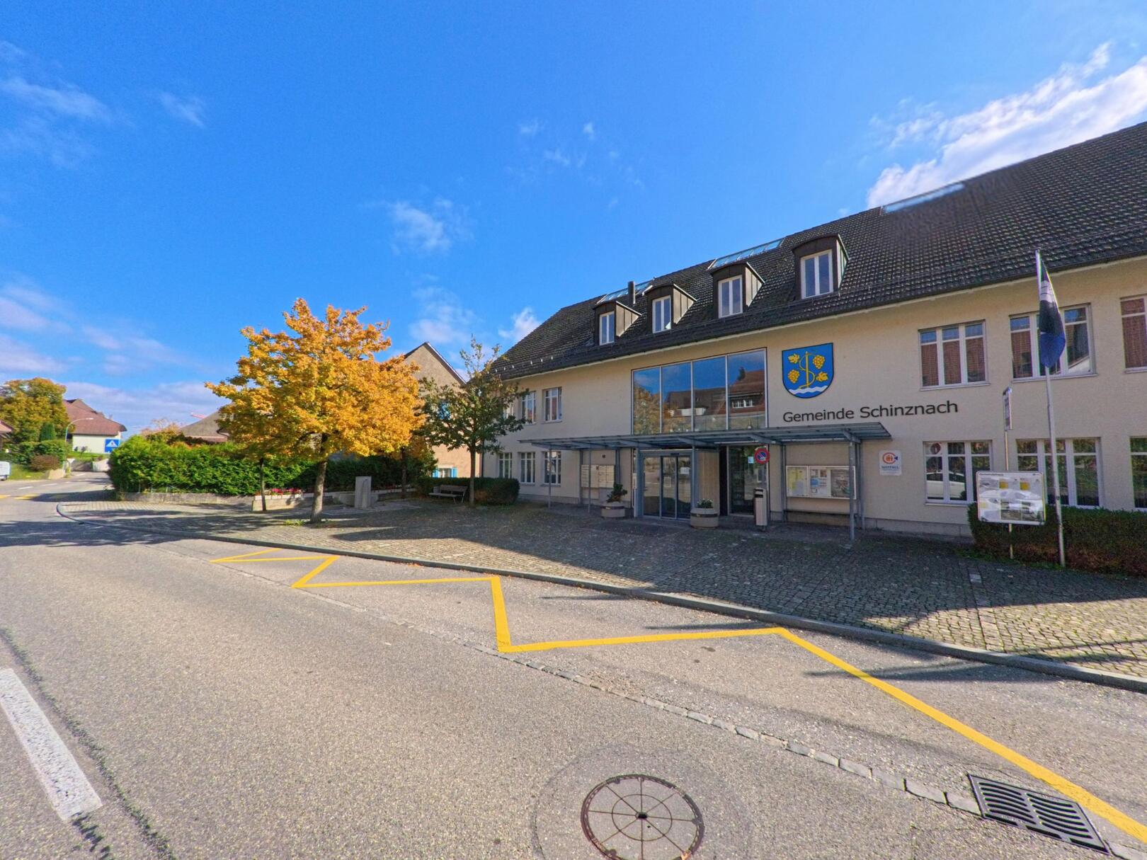 Das Gemeindehaus von Schinznach (5107, Kanton Aargau) steht an einer sonnigen Strasse, vor dem Gebäude wehen Fahnen und ein Baum zeigt herbstliche Farben.