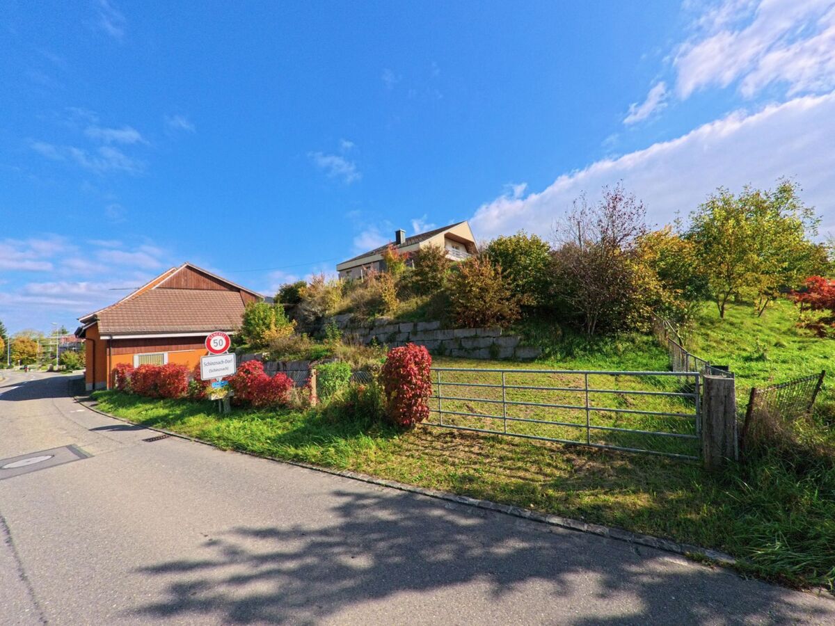 Am Ortseingang von Schinznach (5107, Kanton Aargau) steht ein Schild mit Geschwindigkeitsbegrenzung, daneben Häuser und herbstlich gefärbte Gärten am Hang.
