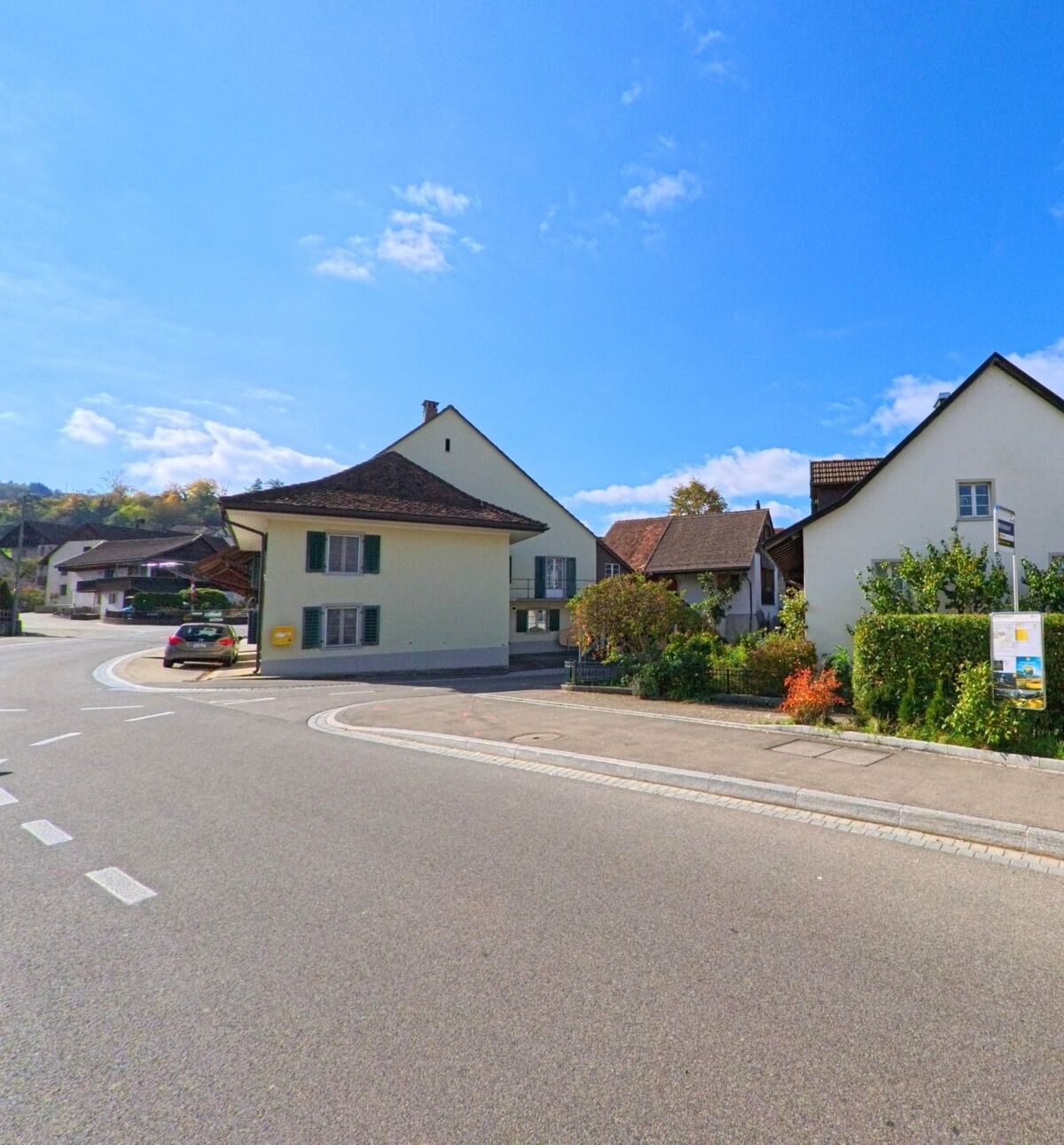 Eine Baumreihe säumt die Landstrasse in Schinznach (5108, Kanton Aargau), dahinter breiten sich grüne Wiesen unter klarem blauen Himmel aus.