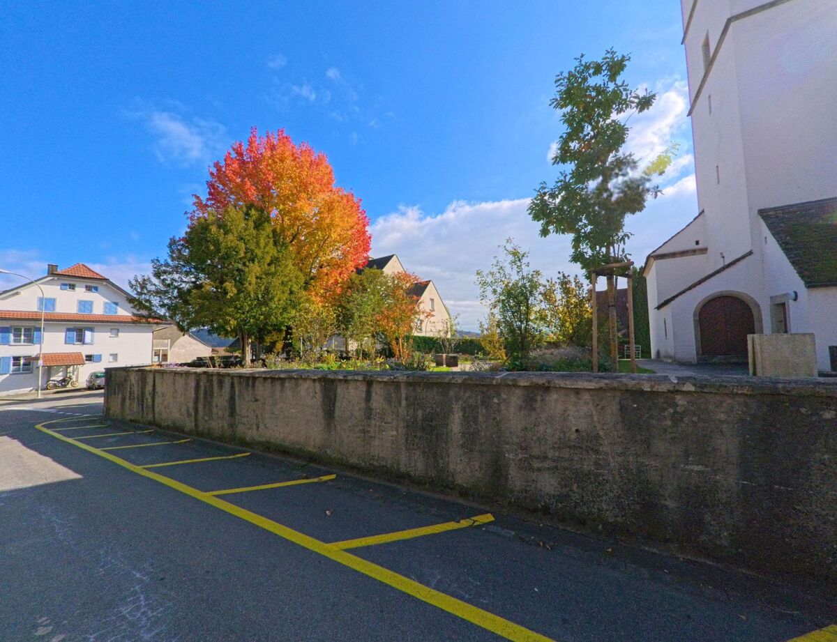 Neben der Kirche in Schinznach (5108, Kanton Aargau) steht ein Baum mit leuchtend rotem Herbstlaub, umgeben von Häusern und einer ruhigen Seitenstrasse.