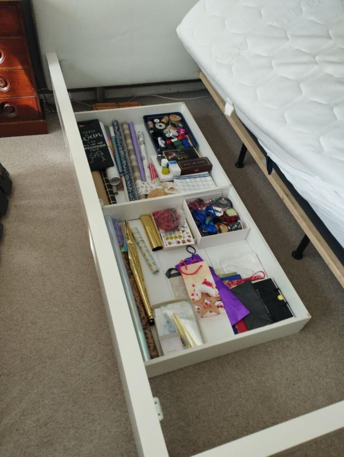 In einem Schlafzimmer in Schlatt (8488, Kanton Zürich) ist eine geöffnete Bettkasten-Schublade zu sehen, die sorgfältig mit Geschenkpapier, Bändern und Bastelmaterial gefüllt ist.