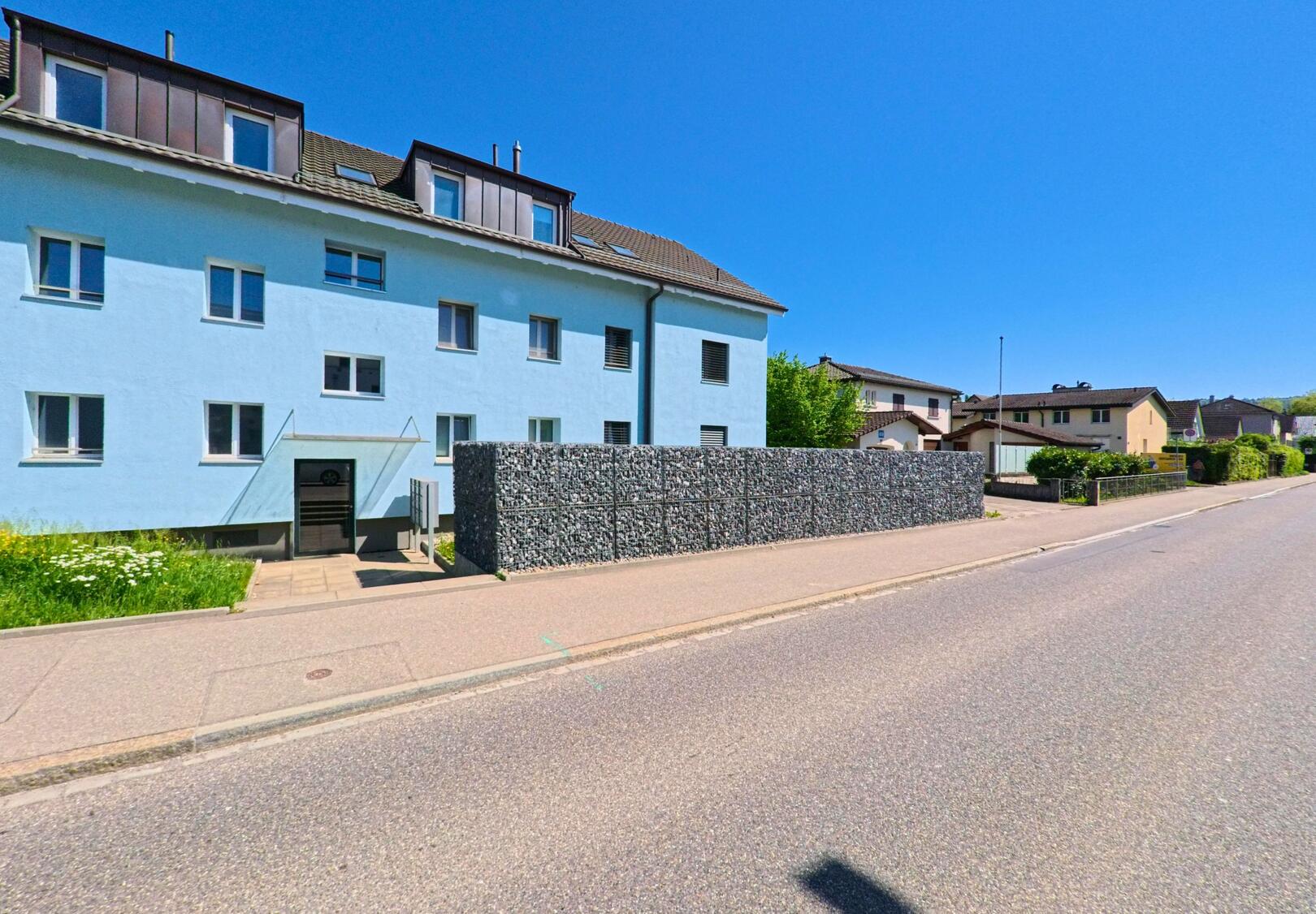 In Schlieren (8952, Kanton Zürich) steht ein hellblaues Mehrfamilienhaus mit moderner Steinmauer entlang einer ruhigen Wohnstrasse.