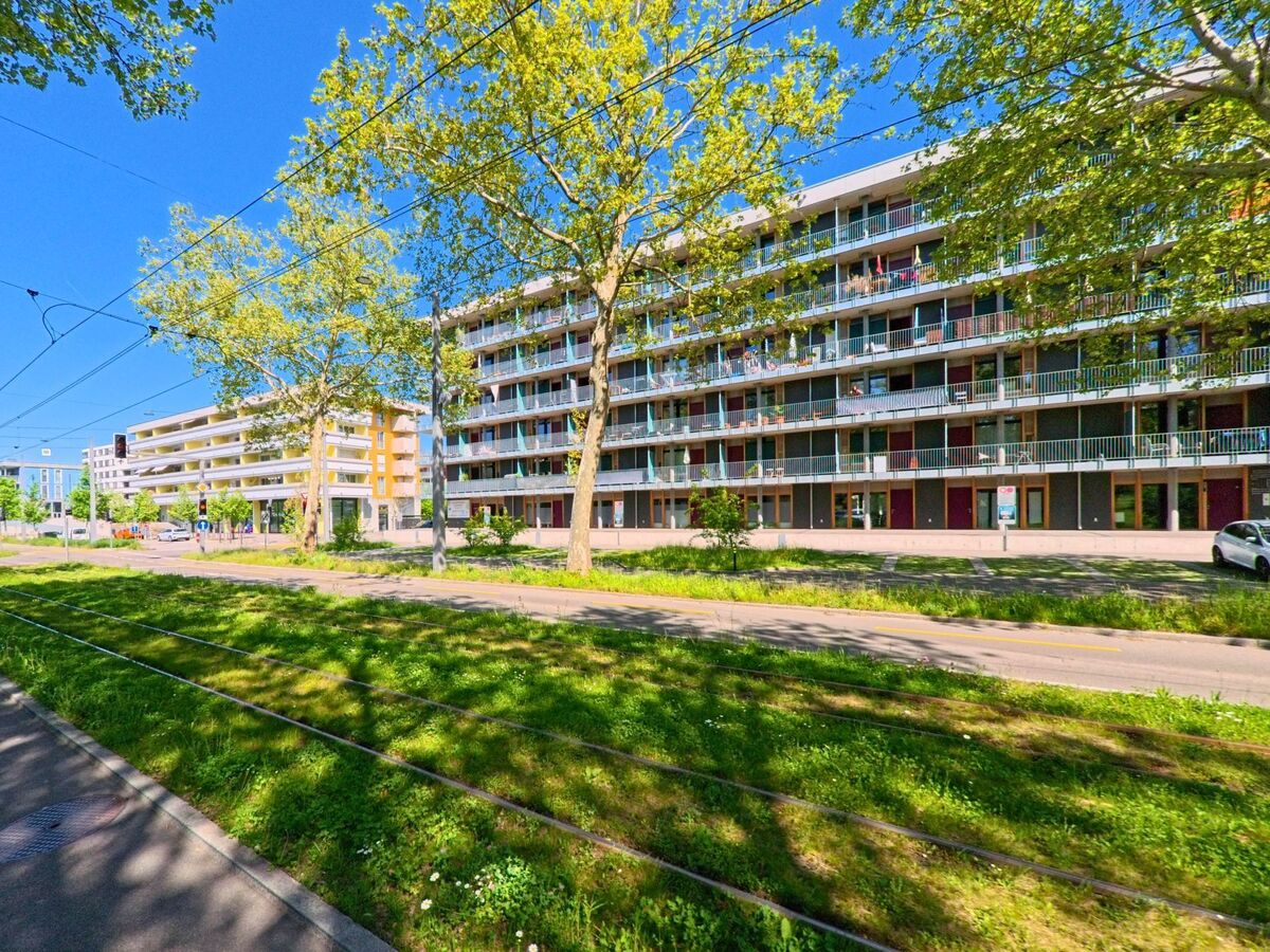 In Schlieren (8952, Kanton Zürich) verlaufen Tramgleise entlang einer Allee mit grossen Bäumen vor modernen Wohnhäusern mit vielen Balkonen.