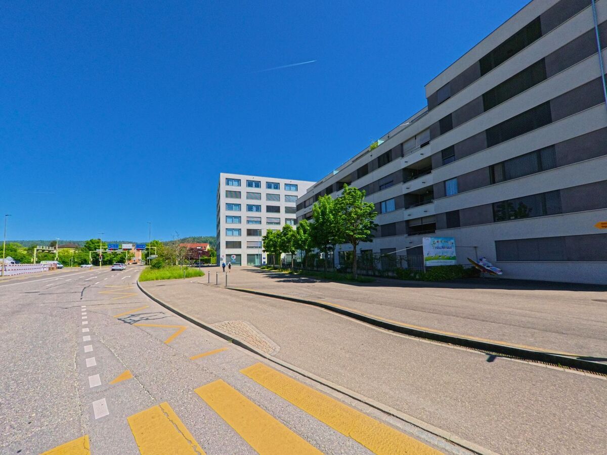 Entlang einer Ausfallstrasse in Schlieren (8952, Kanton Zürich) stehen moderne Wohn- und Geschäftshäuser unter strahlend blauem Himmel.