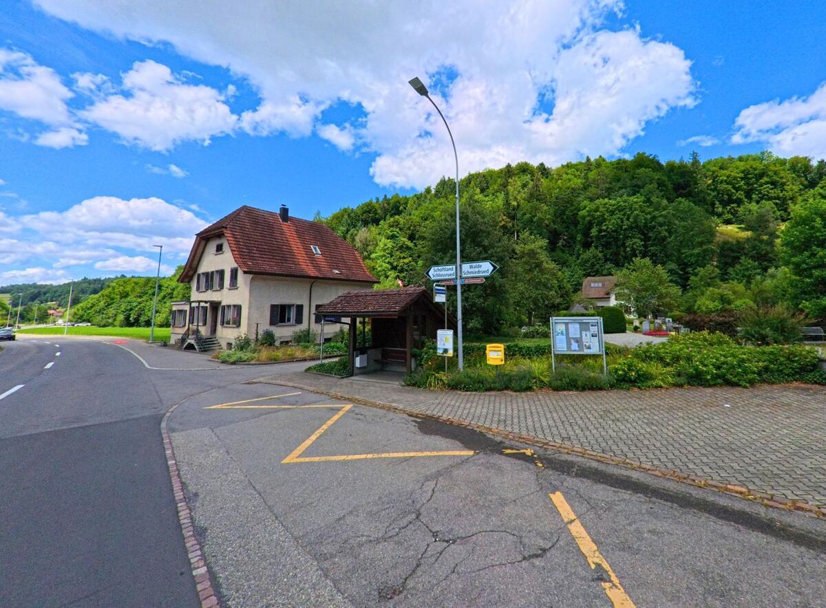 In Schlossrued (5044, Kanton Aargau) liegt eine Bushaltestelle mit Wegweisern und Anschlagtafel an einer Strassenkreuzung neben einem Wohnhaus und vor einem bewaldeten Hang.
