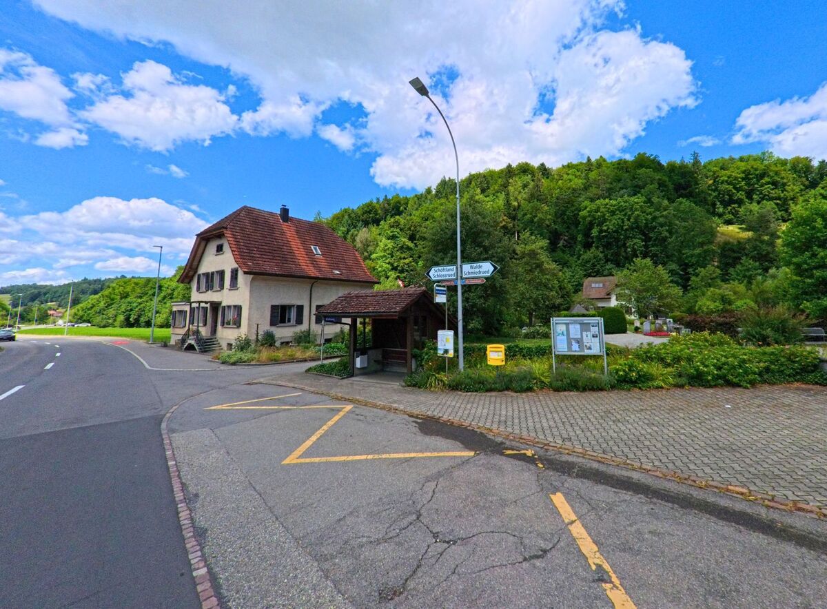 In Schlossrued (5044, Kanton Aargau) liegt eine Bushaltestelle mit Wegweisern und Anschlagtafel an einer Strassenkreuzung neben einem Wohnhaus und vor einem bewaldeten Hang.