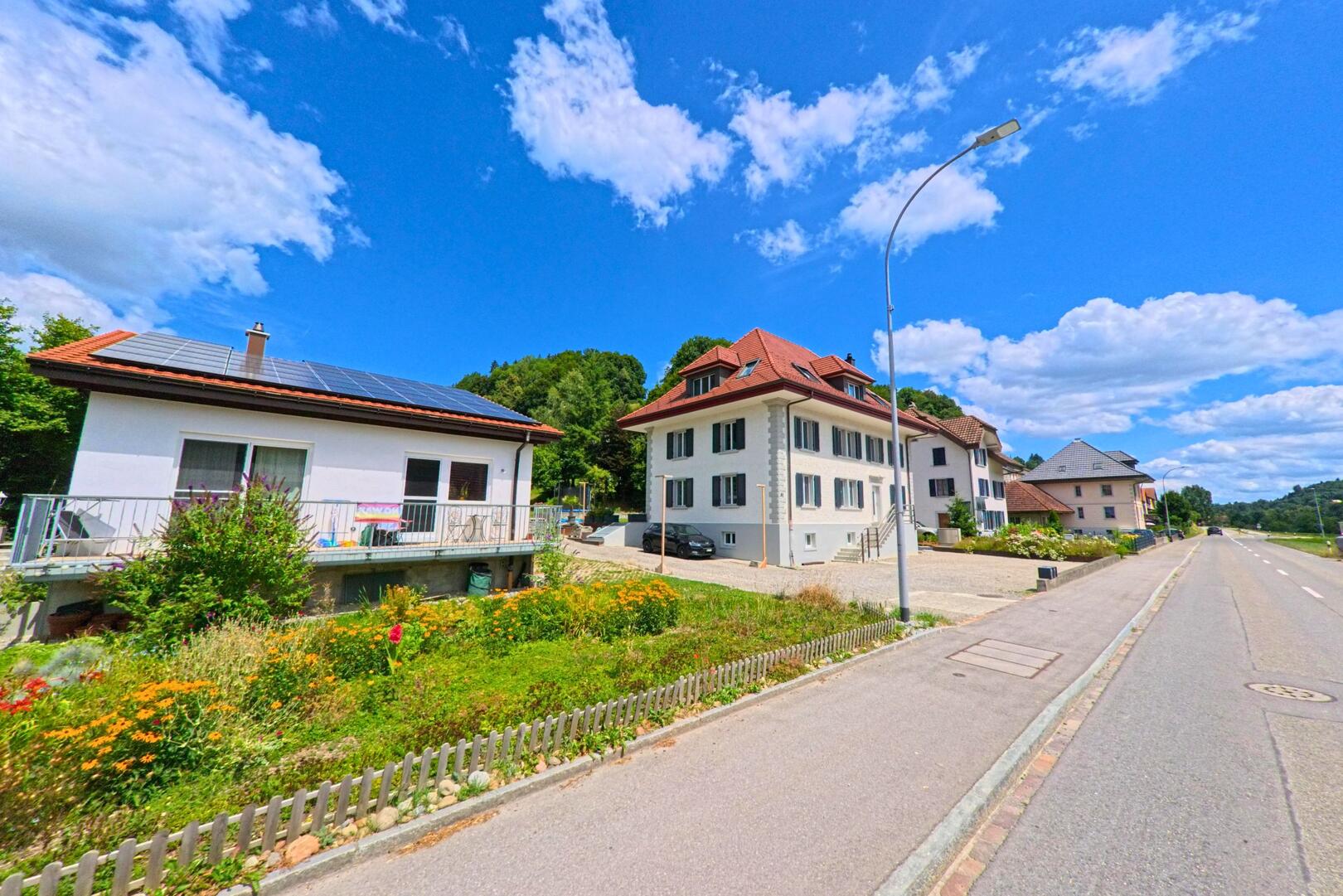 In Schlossrued (5044, Kanton Aargau) stehen mehrere Einfamilienhaeuser mit gepflegten Vorgaerten entlang einer ruhig verlaufenden Strasse unter blauem Himmel.