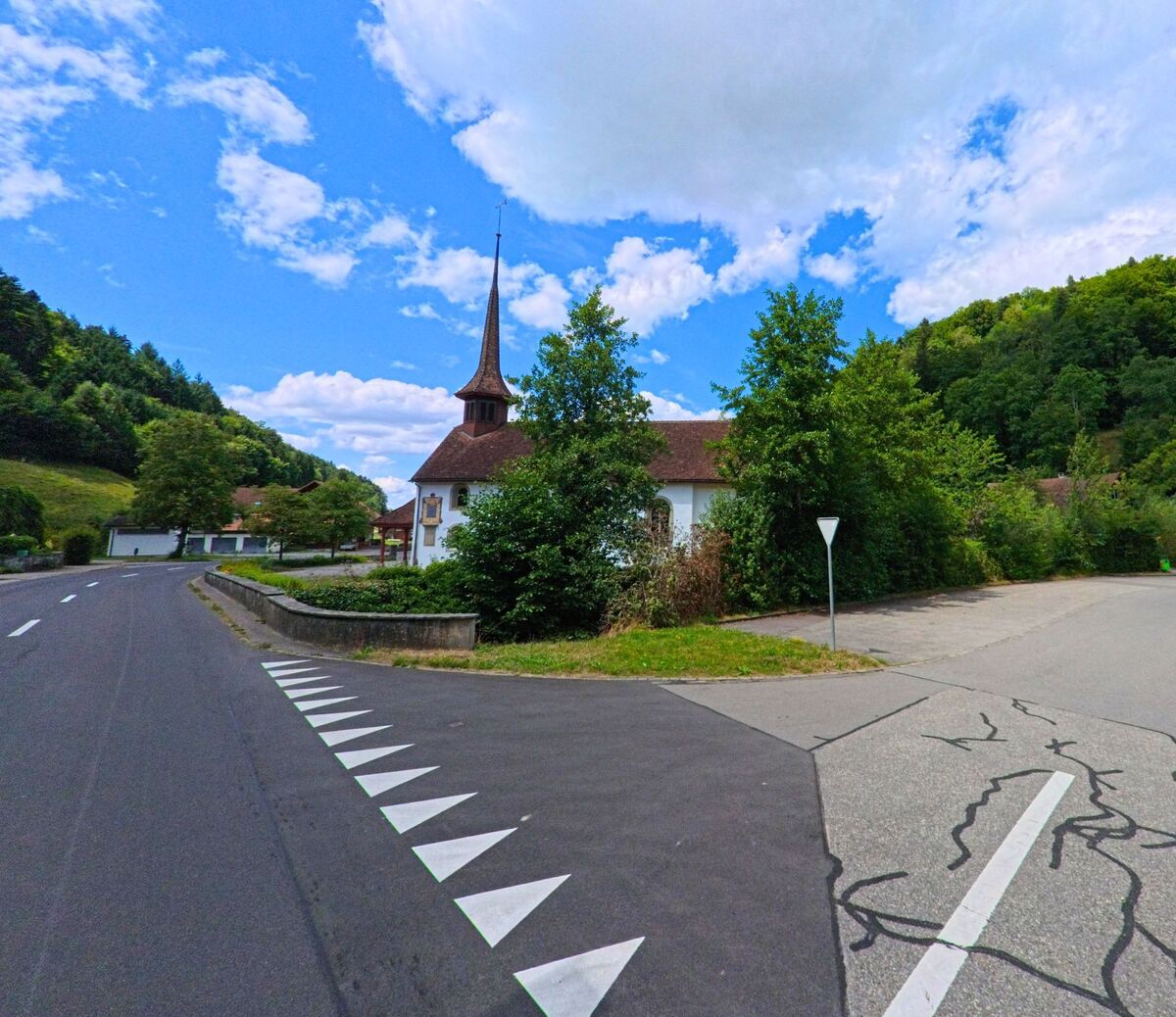 Eine Kirche mit spitzem Turm steht in Schlossrued (5044, Kanton Aargau) an einer Weggabelung, umgeben von Gruenflaechen und bewaldeten Huegeln.