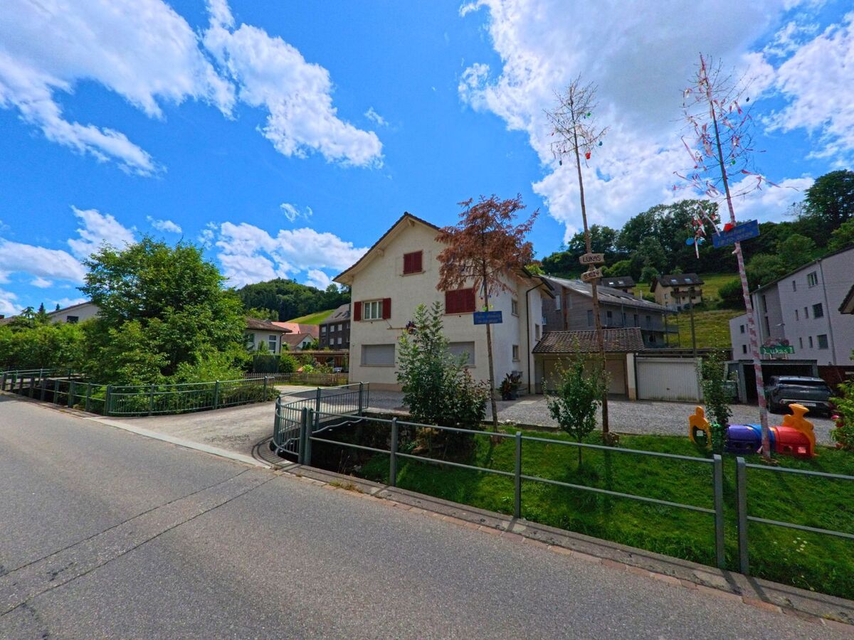 In Schlossrued (5044, Kanton Aargau) markieren bunt geschmueckte Maibaeume vor einem Wohnhaus eine kleine Gruenflaeche mit Spielgeraeten an einer ruhigen Quartierstrasse.