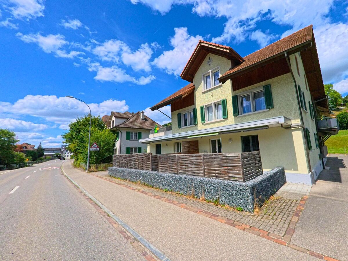 Entlang einer leicht geschwungenen Strasse in Schlossrued (5044, Kanton Aargau) stehen mehrgeschossige Wohnhaeuser mit farbigen Fensterlaeden und einem Hinweis auf einen Schulweg.