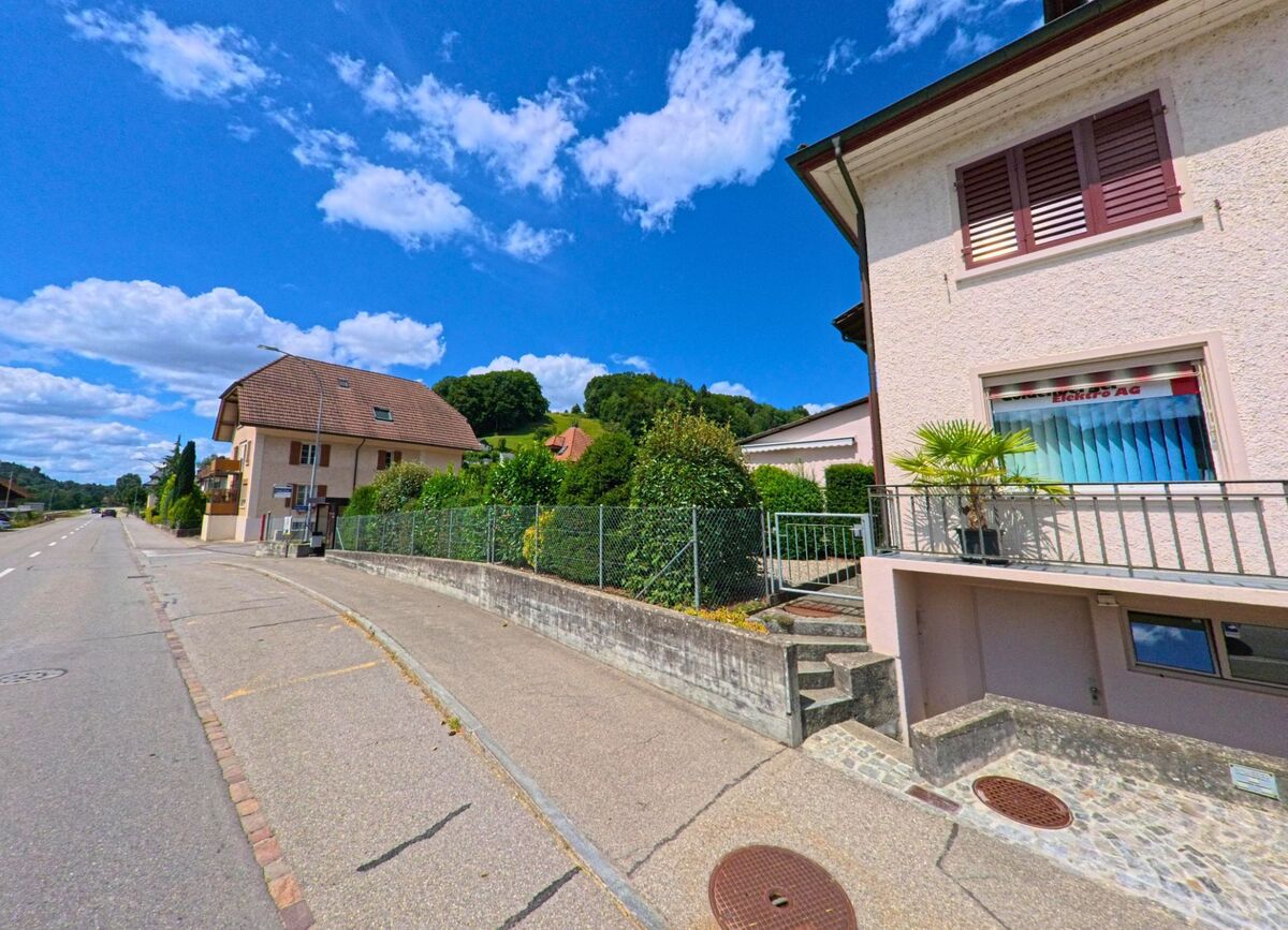 Entlang der Hauptstrasse in Schlossrued (5044, Kanton Aargau) reihen sich Wohnhaeuser mit gepflegten Gaerten aneinander, waehrend der Trottoirbereich leicht ansteigt.
