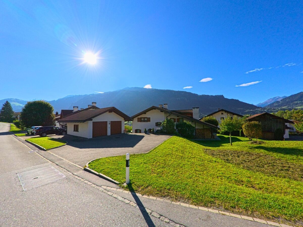 Unter strahlender Sonne liegen in Schluein (7151, Kanton Graubuenden) mehrere Einfamilienhaeuser mit gepflegten Gaerten vor einer eindrucksvollen Bergkulisse.
