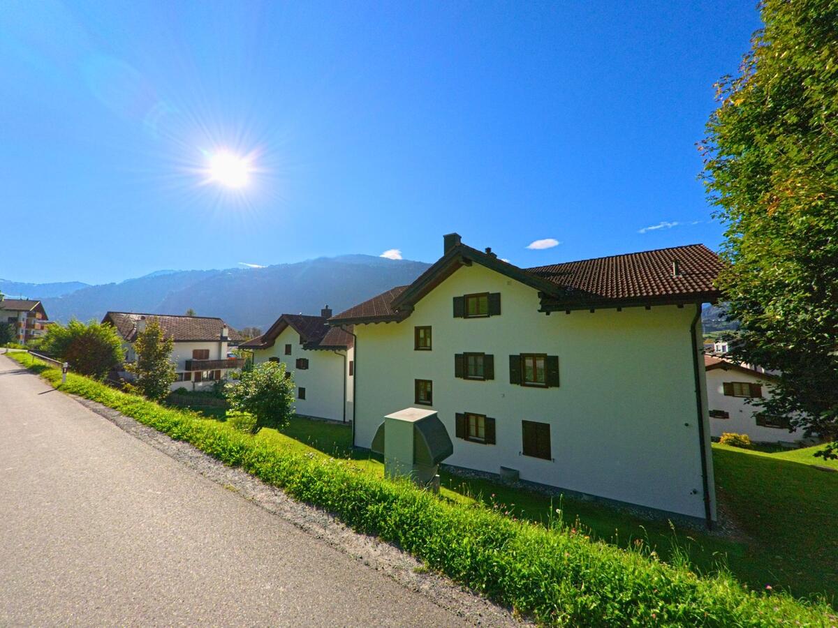 Mehrfamilienhaeuser mit dunklen Fensterlaeden stehen in Schluein (7151, Kanton Graubuenden) an einer ruhigen Strasse mit Blick auf die sonnigen Berge.