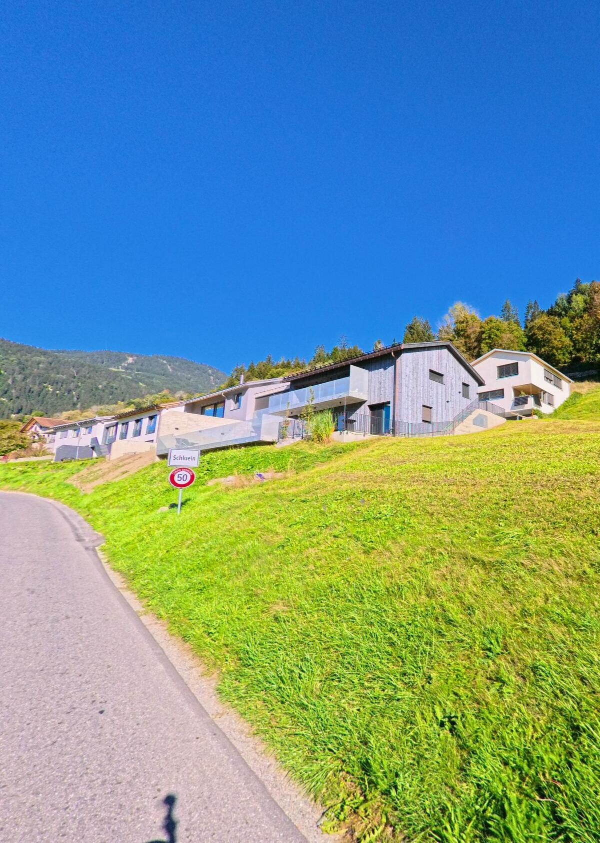 Am Ortseingang von Schluein (7151, Kanton Graubuenden) stehen moderne Wohnhaeuser an einem sonnigen Hang oberhalb der Strasse mit dem Ortsschild.