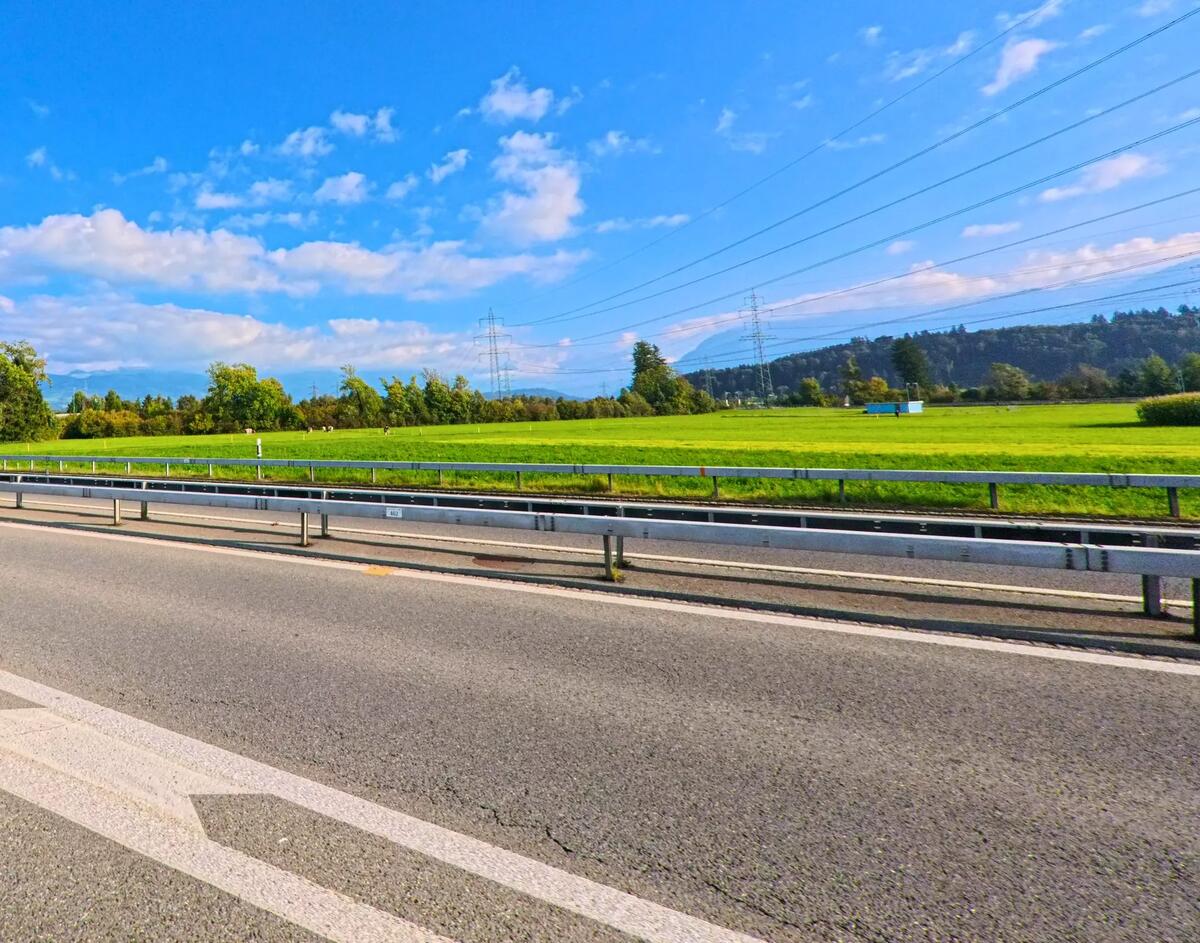 Entlang einer Strasse mit Leitplanke erstreckt sich bei Schmerikon (8716, Kanton St. Gallen) eine weite Gruenflaeche mit Feldern unter blauem Himmel und Stromleitungen.