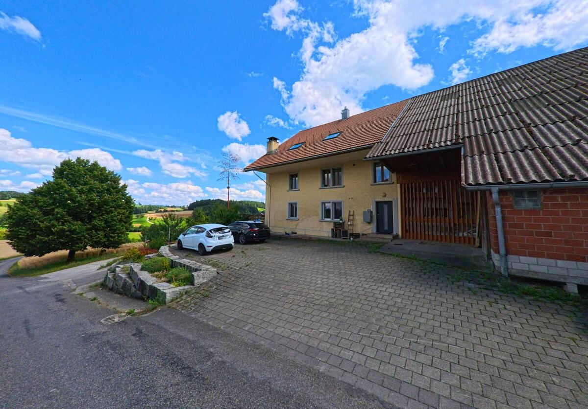 Ein Bauernhaus mit angebautem Wirtschaftsgebaeude steht in Schmiedrued (5046, Kanton Aargau) auf einer Pflasterflaeche oberhalb eines Hangs mit Blick auf die umliegende Felderlandschaft.