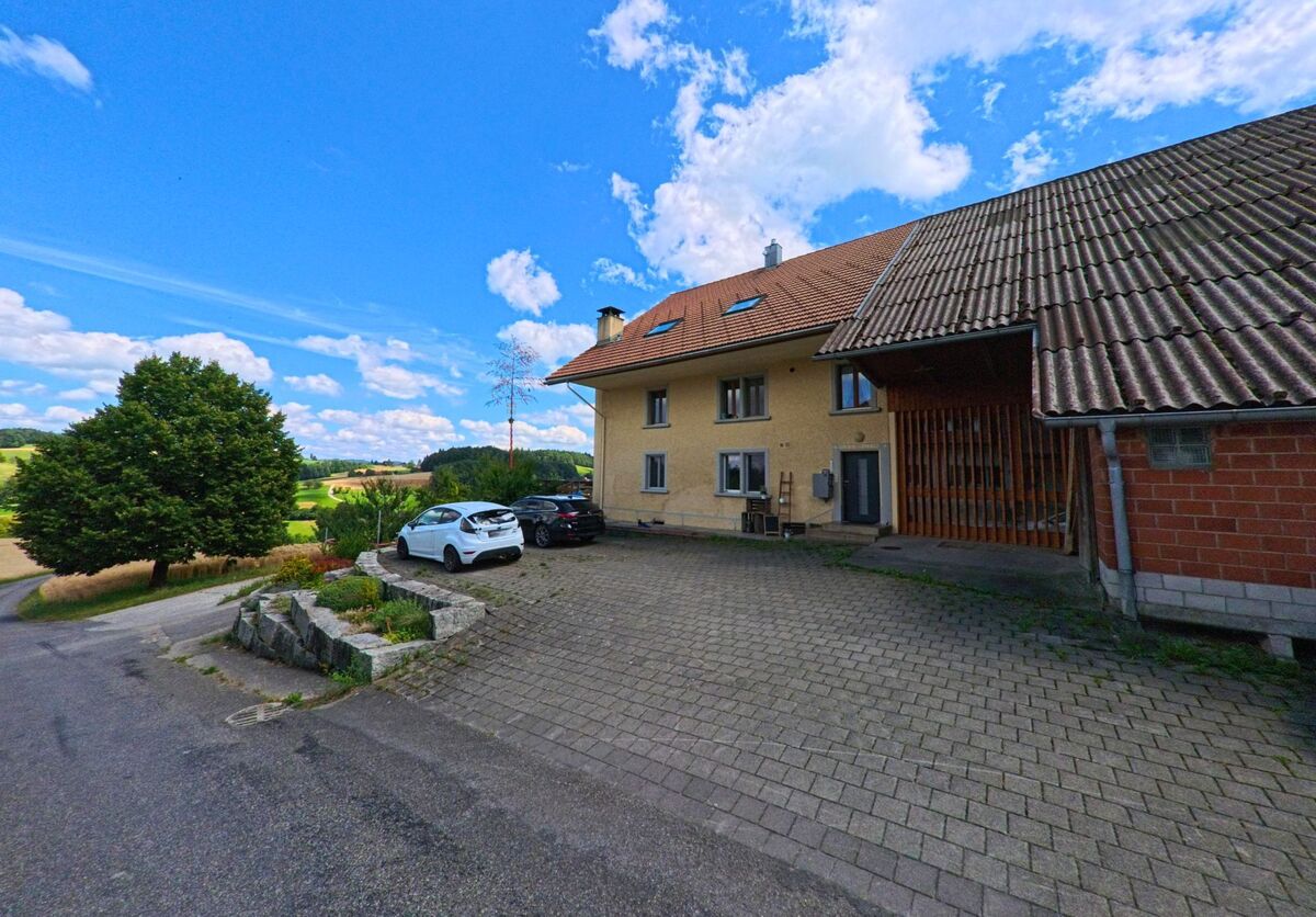 Ein Bauernhaus mit angebautem Wirtschaftsgebaeude steht in Schmiedrued (5046, Kanton Aargau) auf einer Pflasterflaeche oberhalb eines Hangs mit Blick auf die umliegende Felderlandschaft.