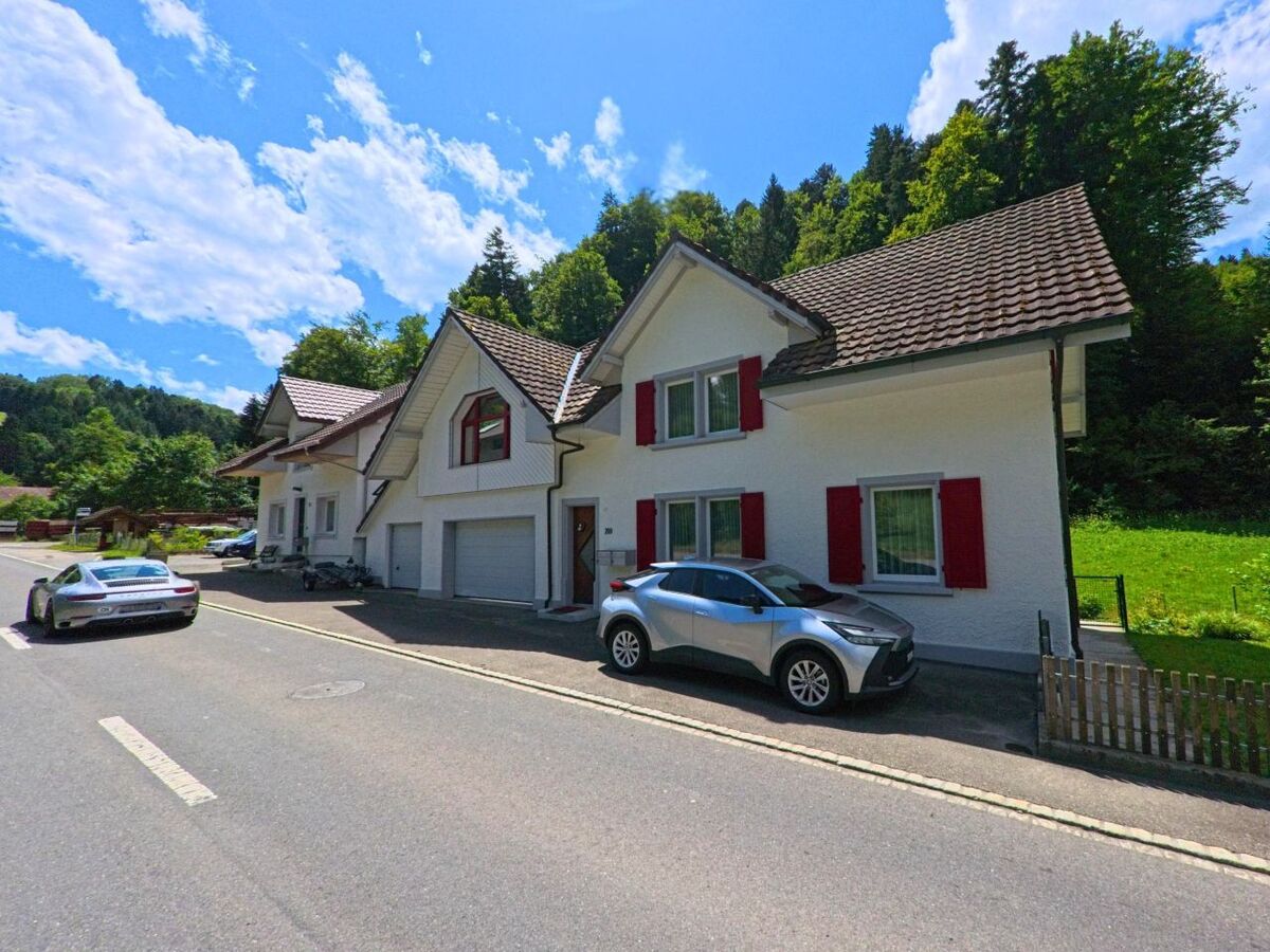 In Schmiedrued (5046, Kanton Aargau) stehen zwei Einfamilienhaeuser mit roten Fensterlaeden direkt an einer Landstrasse, dahinter beginnt ein dichter Waldhang.