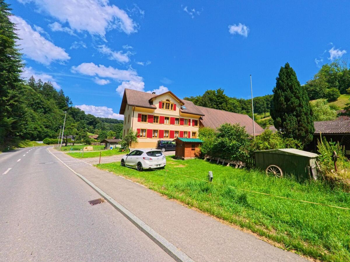 Direkt an der Hauptstrasse von Schmiedrued (5046, Kanton Aargau) steht ein mehrstöckiges Haus mit roten Fensterlaeden und grosszuegigem Gelaende, umgeben von Wald und Huegeln.