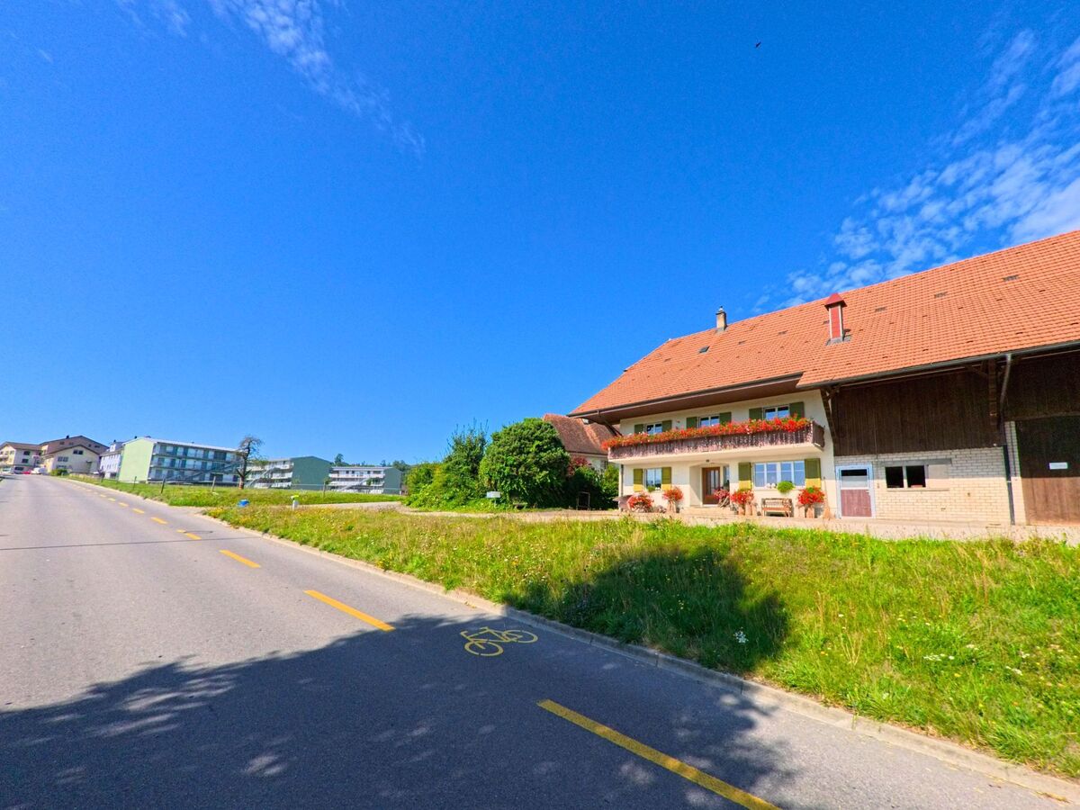 Ein Bauernhaus mit Blumenschmuck am Balkon steht in Schmitten (3185, Kanton Freiburg) an einer Strasse mit markiertem Radweg.