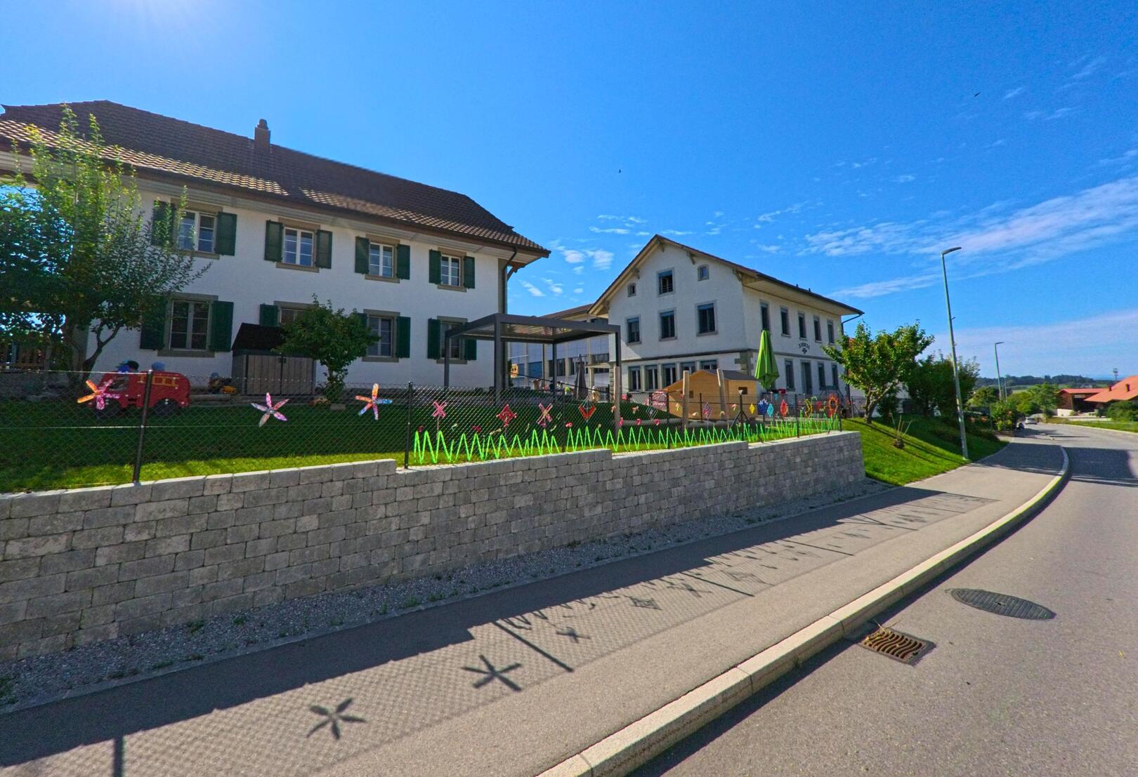 In Schmitten (3185, Kanton Freiburg) steht ein Gebäude mit grün-weissen Fensterläden neben einem umzäunten Spielbereich, der mit bunten Dekorationen geschmückt ist.