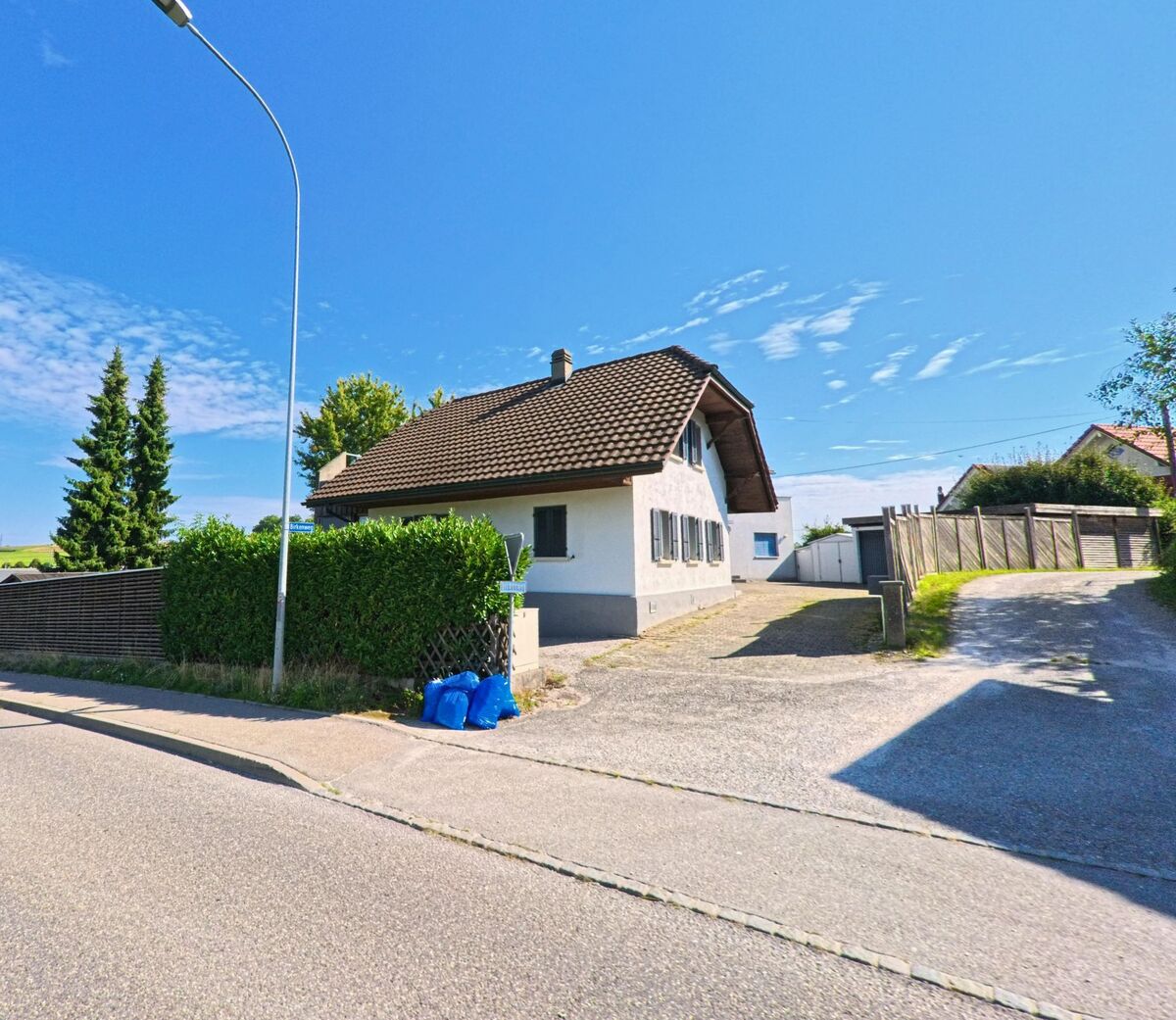 Ein Haus mit braunem Ziegeldach und dunklen Fensterläden steht in Schmitten (3185, Kanton Freiburg) an einer Einfahrt mit angrenzender Hecke.