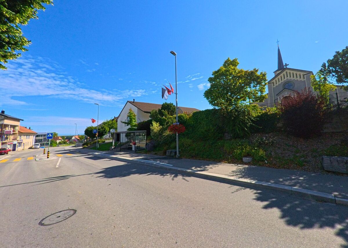 An einer Hauptstrasse in Schmitten (3185, Kanton Freiburg) stehen Wohnhäuser und die Kirche mit ihrem markanten Turm, flankiert von Grünanlagen und Fahnenmasten.