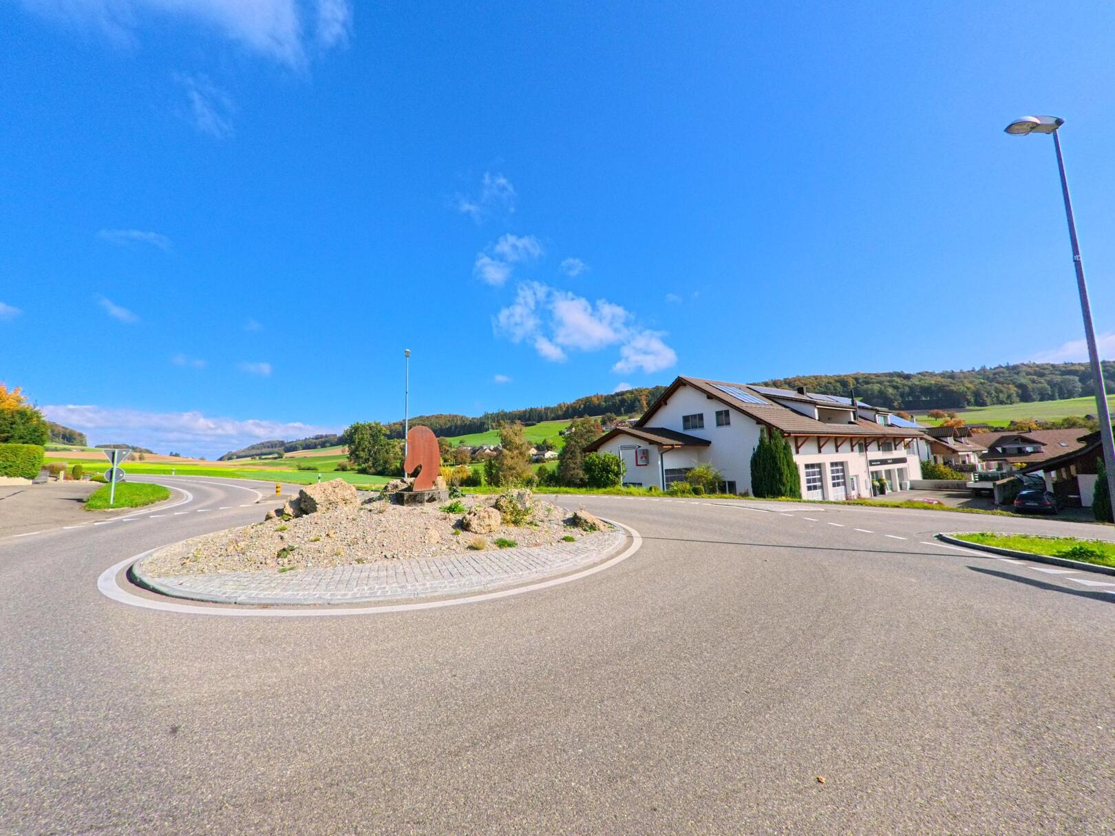 An einem sonnigen Tag zeigt sich in Schneisingen (5425, Kanton Aargau) ein kleiner Kreisel mit einer Metallskulptur, umgeben von Einfamilienhäusern und grünen Hügeln im Hintergrund.
