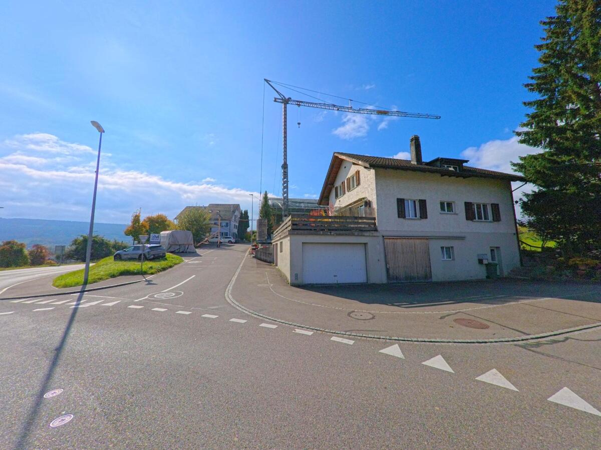 Ein zweigeschossiges Wohnhaus mit geschlossenen Fensterläden steht an einer Kreuzung in Schneisingen (5425, Kanton Aargau), während im Hintergrund ein Baukran zu sehen ist.
