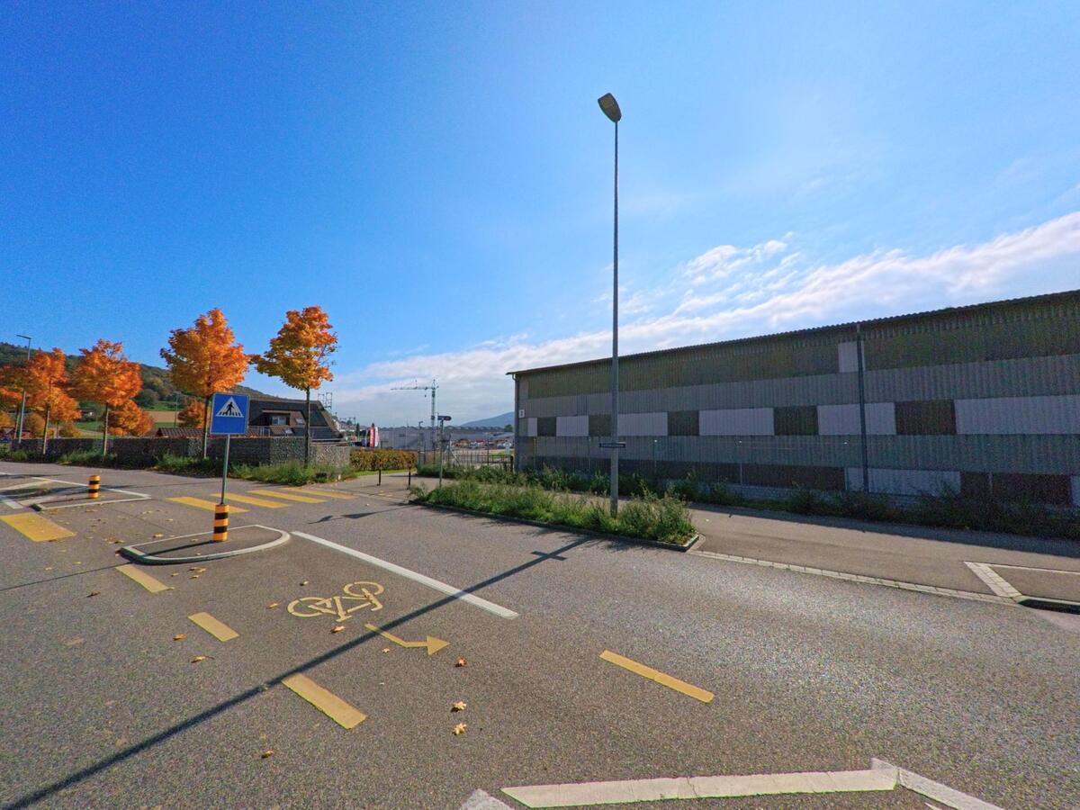In Schneisingen (5425, Kanton Aargau) führt ein Radweg mit Zebrastreifen an einer grossen Industriehalle vorbei, während herbstlich gefärbte Bäume und Baukräne im Hintergrund zu sehen sind.