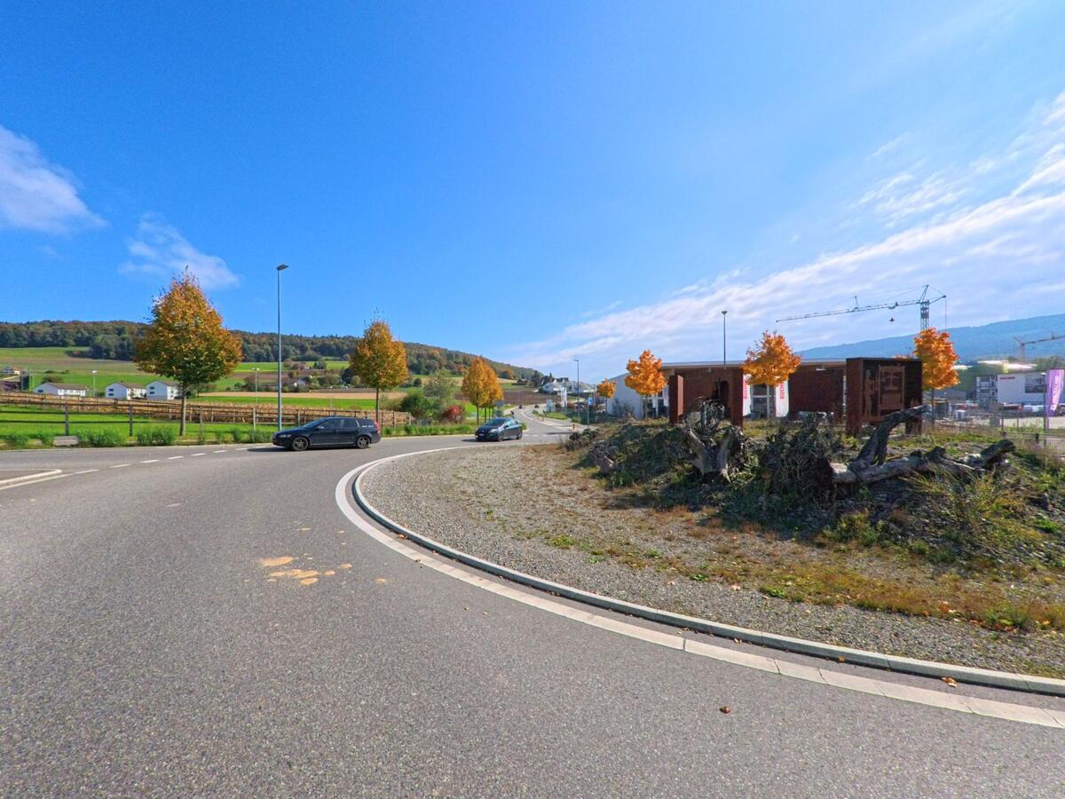Eine Strassenkurve in Schneisingen (5425, Kanton Aargau) führt an jungen Bäumen mit herbstlich gefärbtem Laub vorbei, während im Hintergrund Baukräne und Felder zu sehen sind.