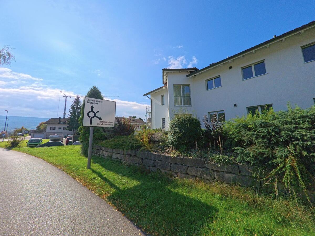 Ein Wegweiser mit Zielangaben nach Zürich, Baden und Dielsdorf steht in Schneisingen (5425, Kanton Aargau) neben einem weissen Wohnhaus unter blauem Himmel.