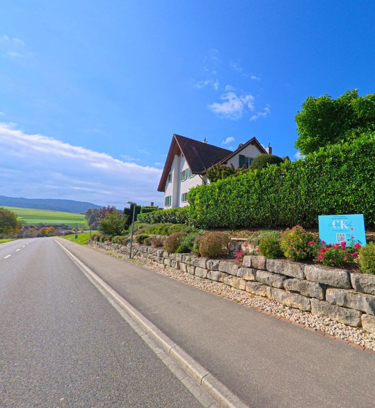 Ein weisses Wohnhaus mit gepflegter Hecke und Blumenbeet steht in Schneisingen (5425, Kanton Aargau) oberhalb einer Landstrasse mit Blick auf das Tal und die Felder.